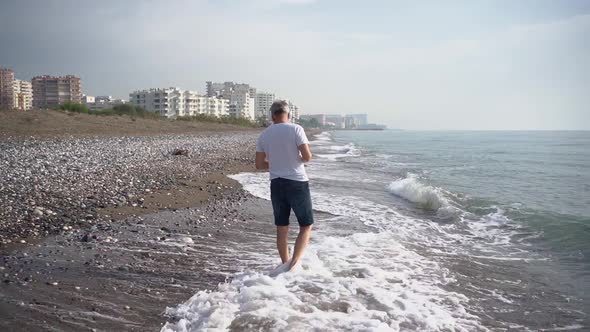 A Slender Elderly Grayhaired Man Slowly Walks Barefoot on a Deserted Beach in the Running Waves in