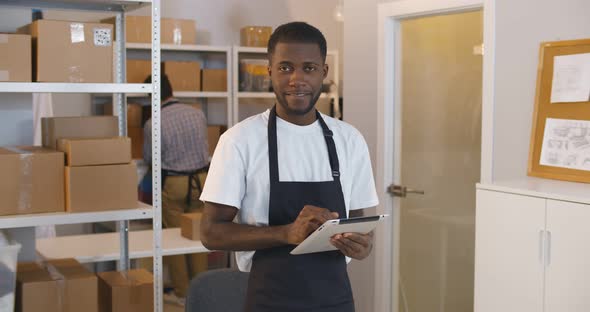 Portrait of Young Mixedrace Man Using Digital Tablet in Warehouse Doing Inventory Smiling at Camera alt