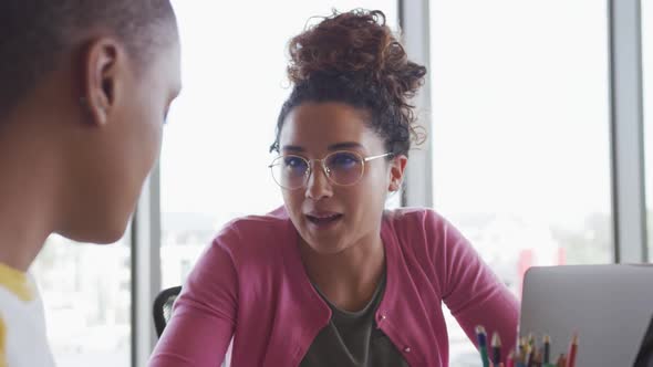 Two happy diverse creative businesswomen using laptops talking at desk in modern interior alt