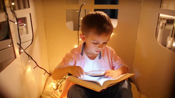 Portrait of Little Boy Holding Glashlight and Magnifying Glass While Reading Book at Night alt