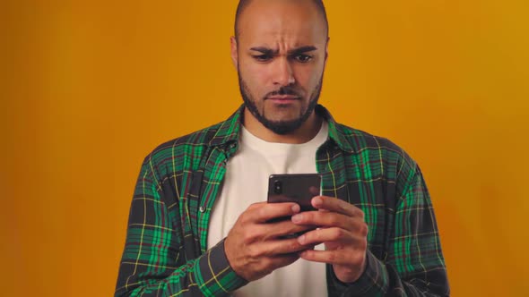 Confused and Surprised African American Man Typing on His Mobile Phone Against Yellow Background alt