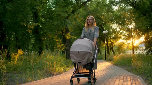 Summer Walk of a Young Beautiful Mother with a Baby Stroller in the Park. Beautiful Mom in a Summer alt