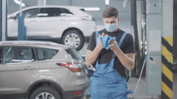Tired Young Caucasian Auto Mechanic in Covid Face Mask Cleaning Hands in Auto Repair Shop alt