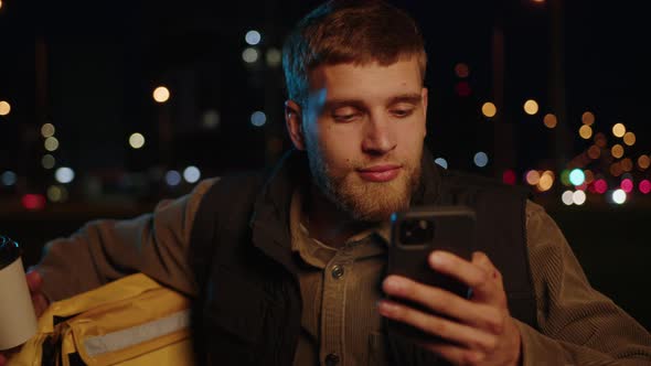 Closeup of a Delivery Man on a Scooter Who Looks at Orders on His Phone in the Evening alt