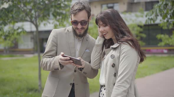 Positive Caucasian Couple Surfing Internet in Smartphone Outdoors. Portrait of Smiling Young Man and alt