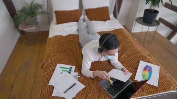 Top View Wide Shot of Young Asian Man Lying on Bed in Headphones and Covid19 Face Mask Messaging alt