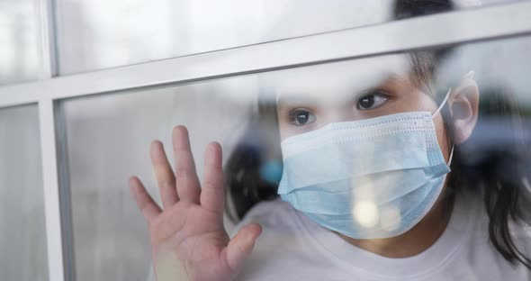 little Asian girl in face mask looking out window alt