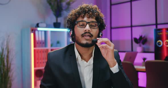 Indian Businessman in Headset Talking Into Camera on Evening Office Room Background alt