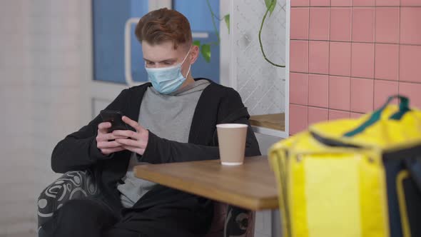 Young Caucasian Man in Covid Face Mask Surfing Internet on Smartphone Waiting for Take Away Order in alt