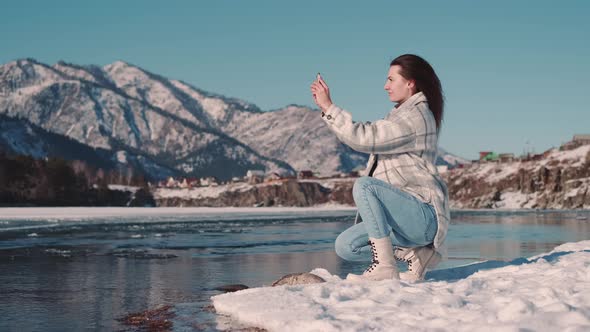 A Woman Tourist Blogger Sits on the River Bank in a Mountainous Area and Takes a Photo Using a alt