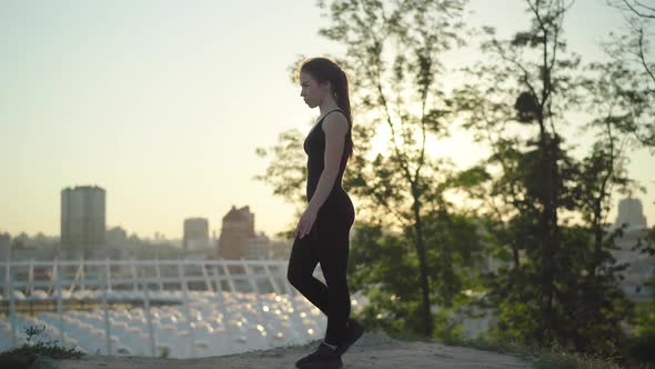 Wide Shot of Confident Caucasian Female Gymnast Walking at Sunset Outdoors and Jumping Up. Side View alt