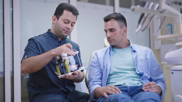 Concentrated Caucasian Man Listening Male Middle Eastern Dentist Talking Showing Dental Cast alt