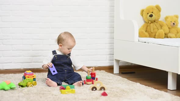 Baby Boy in Denim Overalls Plays with Toys Sitting on the Carpet alt