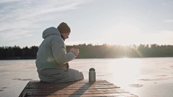 Man Drinks Hot Tea From Thermos Sitting on a Lake Dock on a Sunny Winter Day