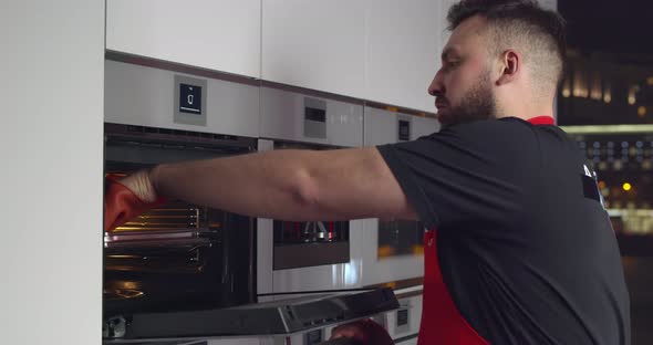 Young Man in Apron Taking Tray with Meal From Electric Oven in Kitchen alt