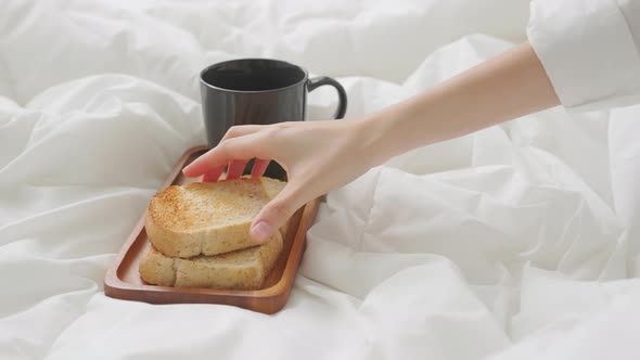 Woman hand eating coffee and bread for breakfast in the bedroom. alt