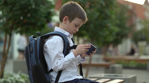 A Schoolboy Sits on a Bench in the Park and Plays on His Phone After Studying at School