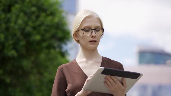 Serious Woman in Glasses with a Tablet on the Background of Skyscrapers