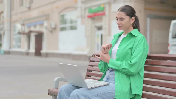 Hispanic Woman with Wrist Pain Using Laptop While Sitting on Bench alt