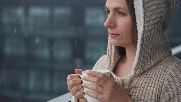 Caucasian Woman Stays on Balcony During Snowfall with Cup of Hot Coffee or Tea alt