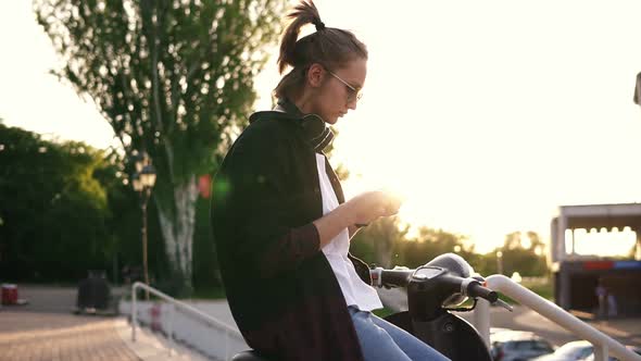 Young Girl in Black Hoodie Sitting on a Motorbike and Typing on Her Mobile alt