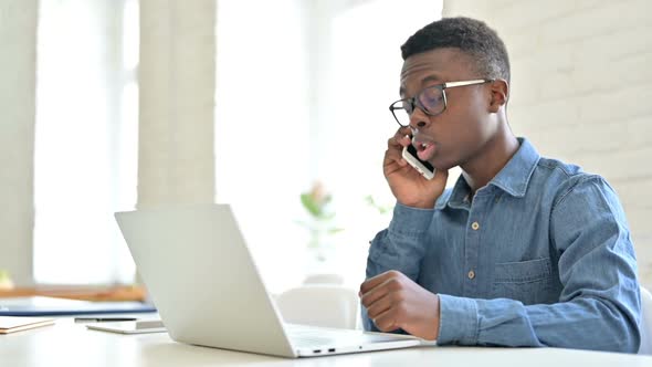 Young African Man with Laptop Talking on Smartphone in Office alt