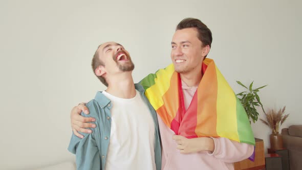 Two Young Men Stand Together Covered with a Lgbt Flag and Talk to Each Other Smiling alt