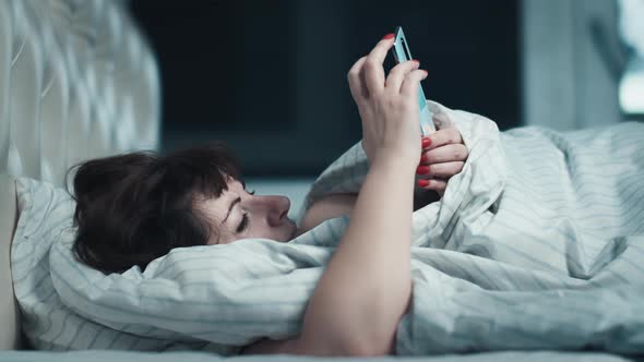 Sleepy Middleaged Woman Lies on the Bed and Uses the Phone at Night