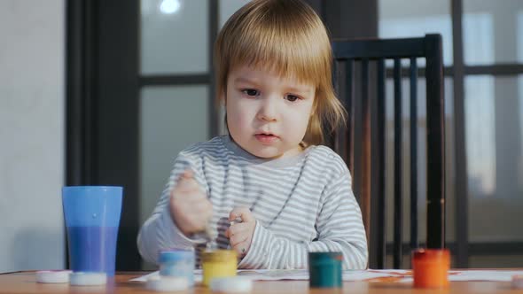 The Kid Painting with Paints and Brush Sitting at the Table