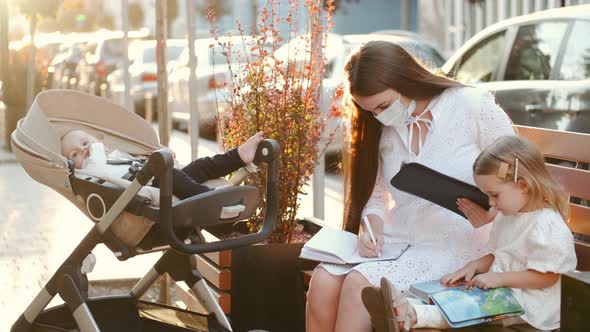 Beautiful Mother in Protective Mask and Two Kids on a Bench in a City alt