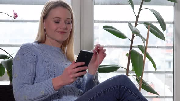 A Young Woman Sits on a Chair By a Window and Listens To Music on a Smartphone with a Smile alt