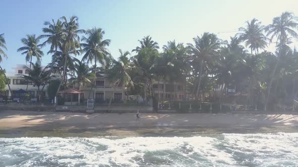 People Walk Along Sand Between Foaming Ocean Waves and Palms alt