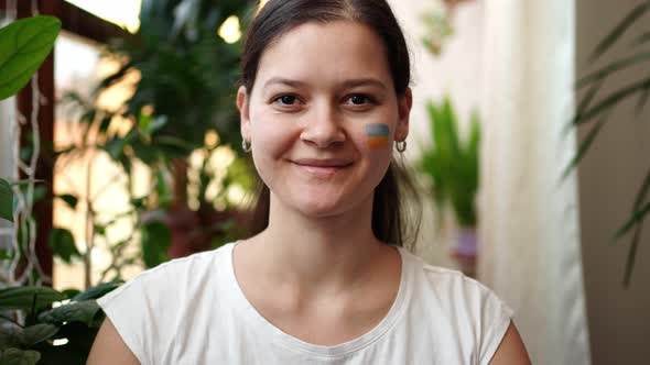 A Smiling Young Ukrainian Girl with the Flag of Ukraine on Her Face is Looking to Camera alt