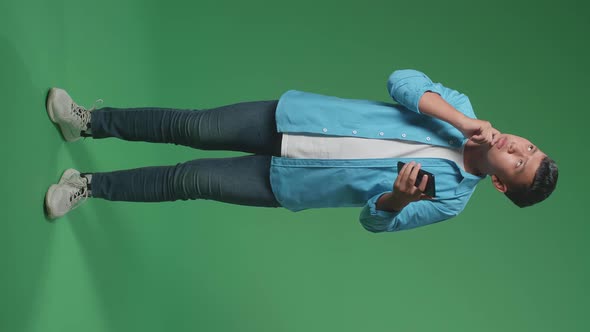 Full Body Of Asian Boy Use Phone And Thinking While Standing On Green Screen In The Studio alt