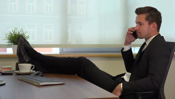 An office worker in a suit sits in front of a computer with legs on desk and talks on a smartphone alt
