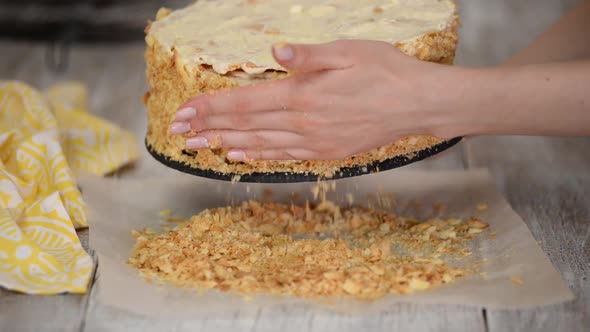 A woman decorates a cake smeared with cream with biscuit crumbs.