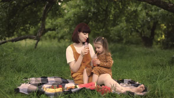 Young Mom with Her Little Cute Daughter on a Picnic in Green Park Drinking Fresh Juice alt