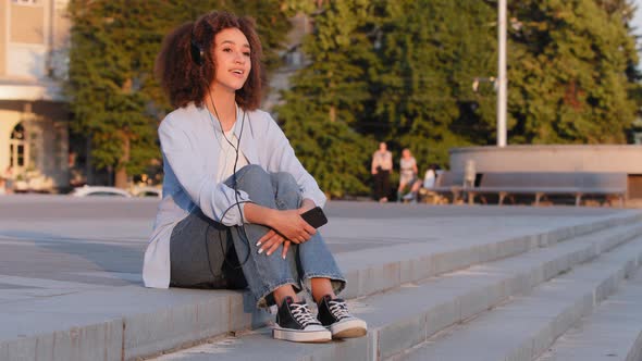 Young Afro American Girl Student African Woman Sitting on Street Outdoors in City Wears Headphones alt