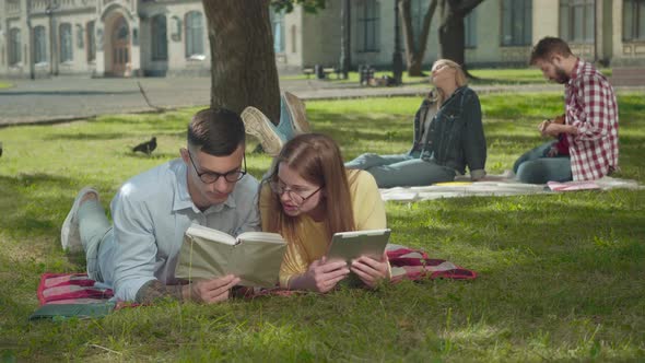 Intelligent Young Boy and Girl in Eyeglasses Lying on Green Lawn and Reading Books As Loving Couple alt