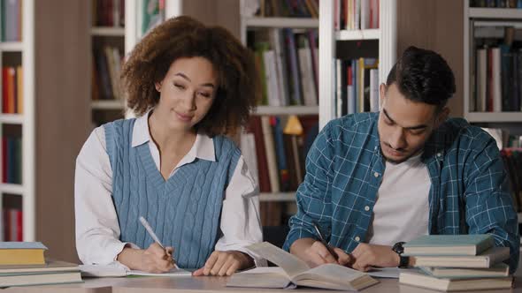 Two Students Young Guy and Girl Doing Homework Sitting at Desk in Library Writing Notes Attentively alt