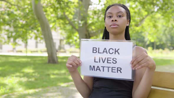 A Young Black Woman Shows a Black Lives Matter Sign To the Camera and Nods As She Sits on a Bench alt