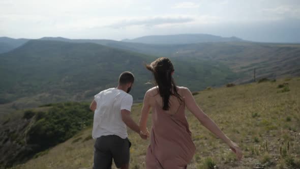 The Happy Young Couple Holding Hands Runs From the Mountain.