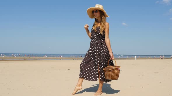 Young Woman Walks On The Beach With Picnic Basket. Fashionable Girl Is Walking On The Beach
