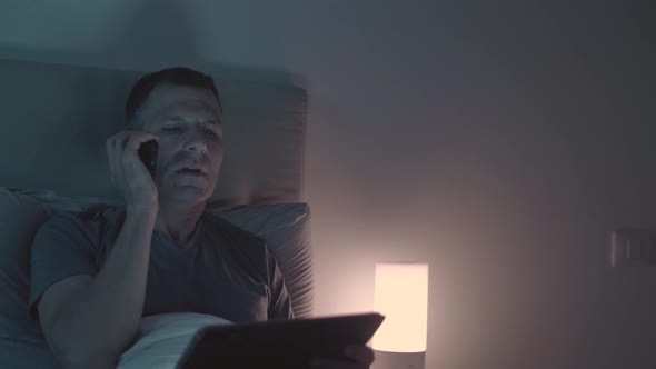 Man Speaking On The Phone And Looking At The Tablet While In Bed At Night