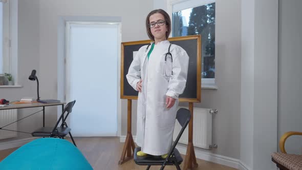 Zoom in to Confident Caucasian Doctor Little Person in Eyeglasses and Medical Gown Crossing Hands alt