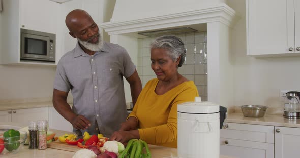 African american senior man hugging his wife while chopping vegetables in the kitchen at home alt