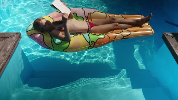 Happy Smart Girl Sunbathing on Inflatable Mattress in Swimming Pool Reading Book in Sunbeam alt