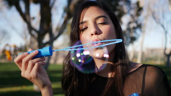 Cute hispanic woman blowing bubbles while smiling and looking happy and nostalgic outdoors in sunlig alt
