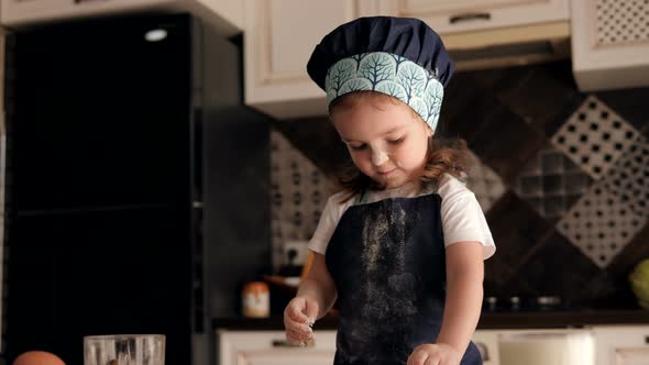 A Little Funny Girl in a Chef's Hat Eats Flour with Her Hands on the Kitchen. alt