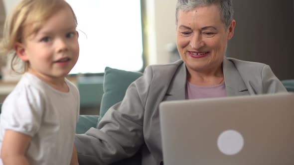 Portrait of a Stylish Grandmother Working at Home on the Couch and Her Little Blondie Grandson alt